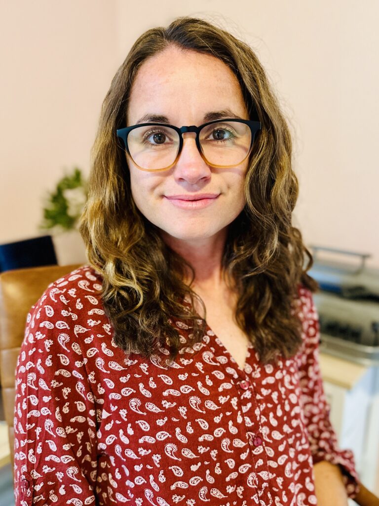 Professional headshot of Genevieve Barnes, sitting in a leather chair, facing the camera, wearing a red blouse and glasses. A laptop and braille writer are on a desk in the background.