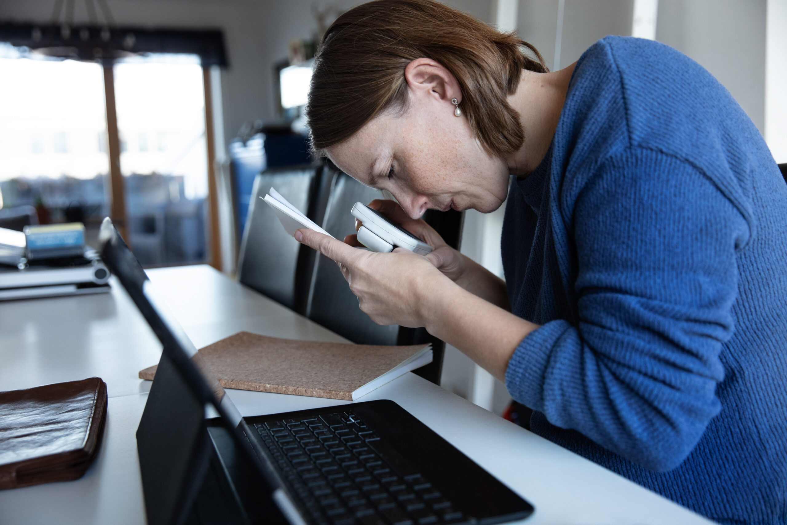 A woman wearing a blue top sitting at a desk in a meeting room with a laptop in front of her holds a magnifier to a smart phone close to her face