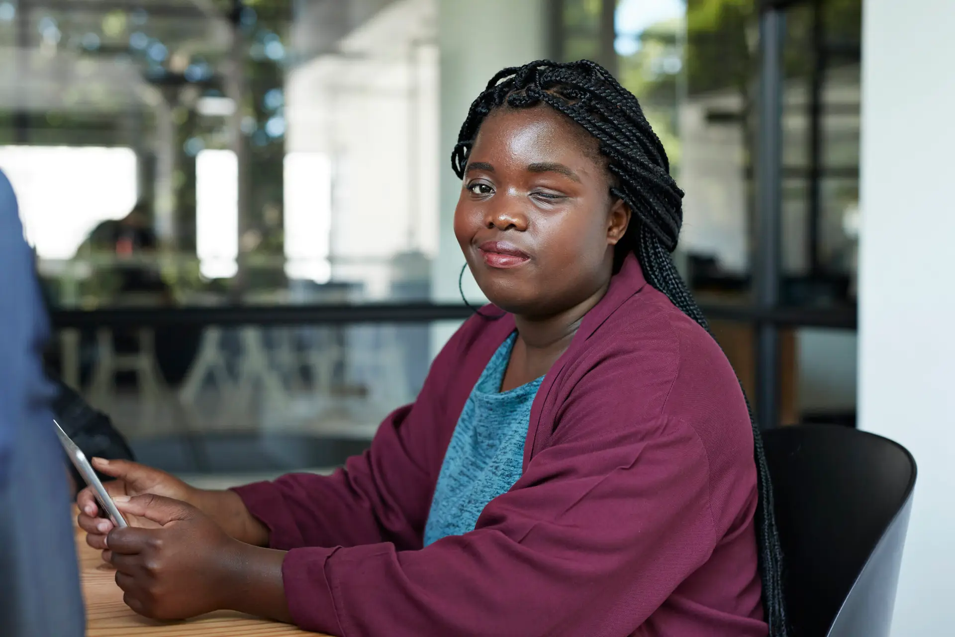 A woman in an office with a visual impairment wearing a blue top and maroon sweater is holding a smart phone and smiling towards the camera