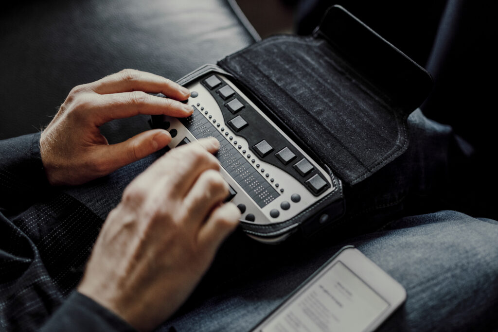 A man's hands reading on a bluetooth braille display. He is wearing blue jeans, sitting on a black leather couch, and a smart phone is rest on his leg with text on screen.