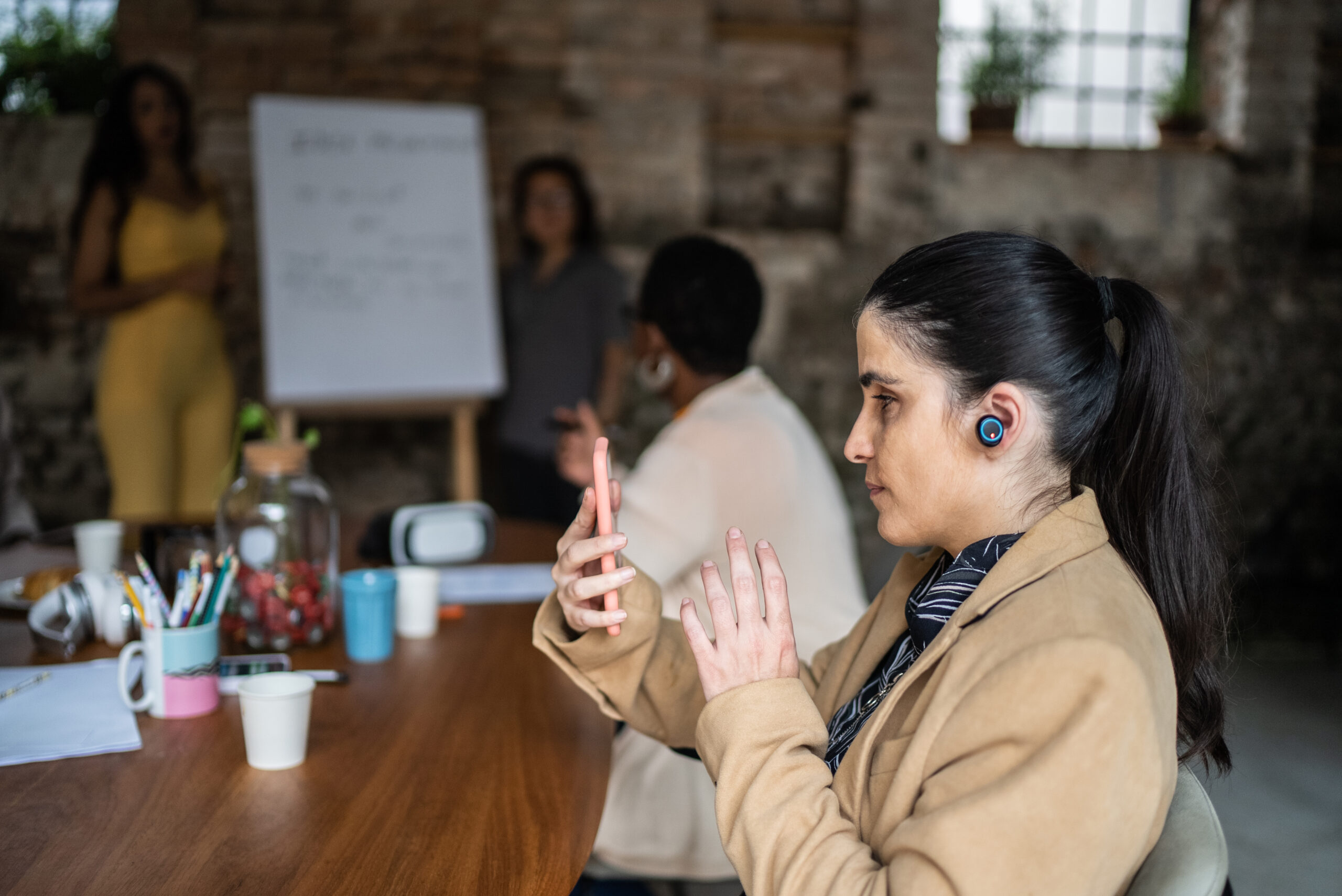 Woman with a visual impairment in a jacket sitting with colleagues at a large table, reads from a smartphone and wears earbuds during a business meeting. A white board with text is in the background.