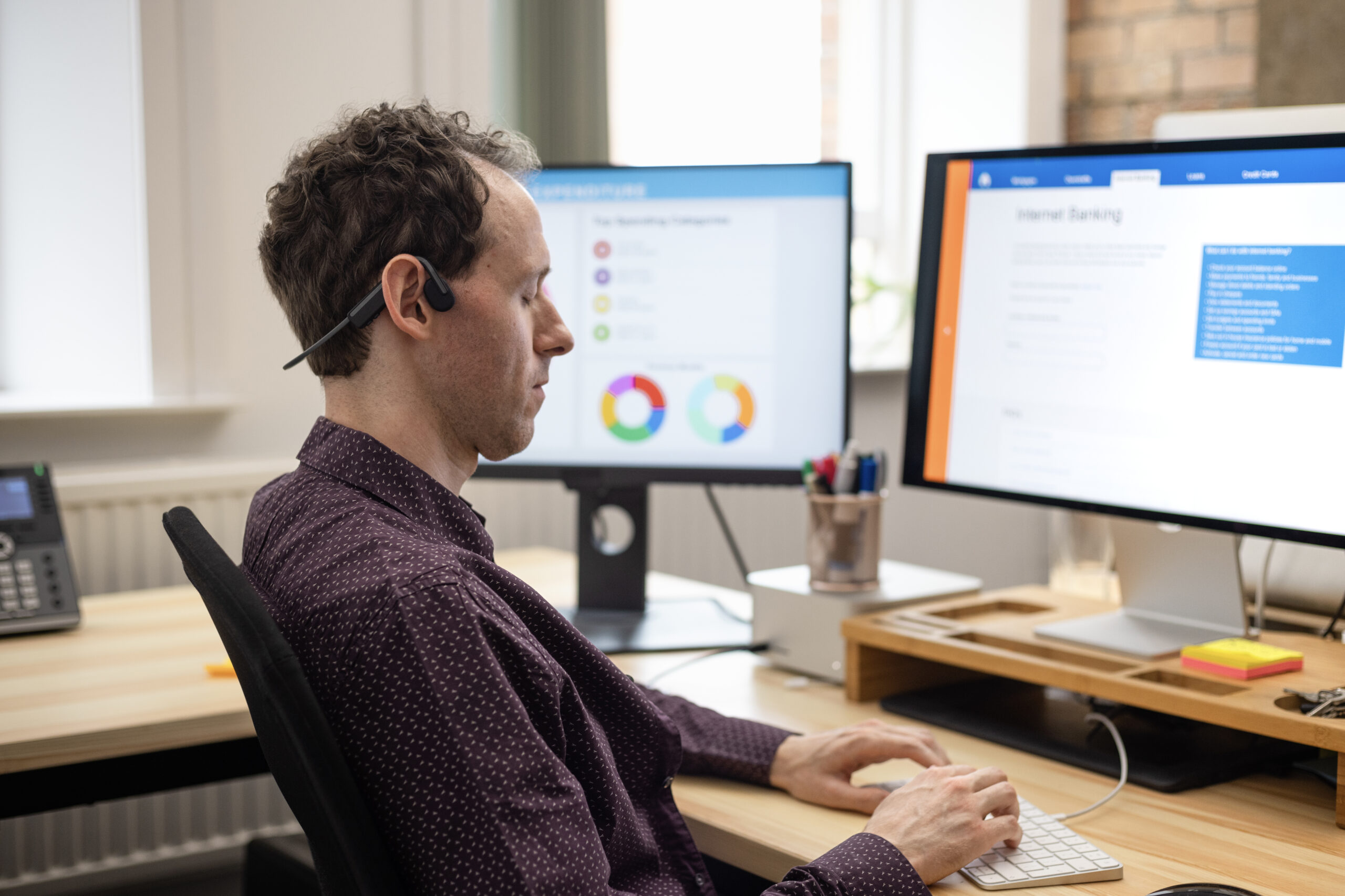 A man with closed eyes wearing a purple shirt and a headset sitting at a desk is facing two computer screens, typing on a keyboard. The screens include graphs and data.