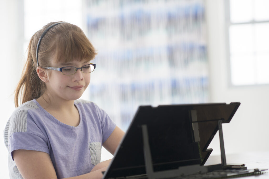 A girl with low vision, wearing a pony tail, a lavender t-shirt and glasses smiling at a tablet mounted on a table.