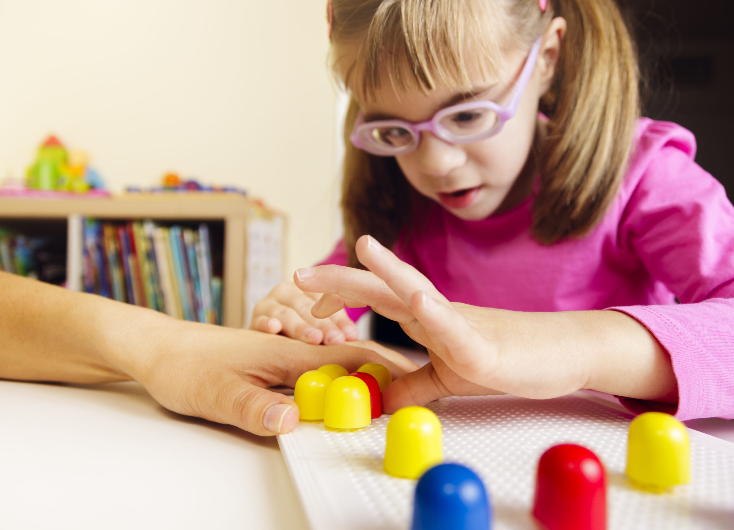 Young girl with low vision wearing a purple top and glasses, reaching for mosaic pegs on the board with an adult hand underneath her hand.