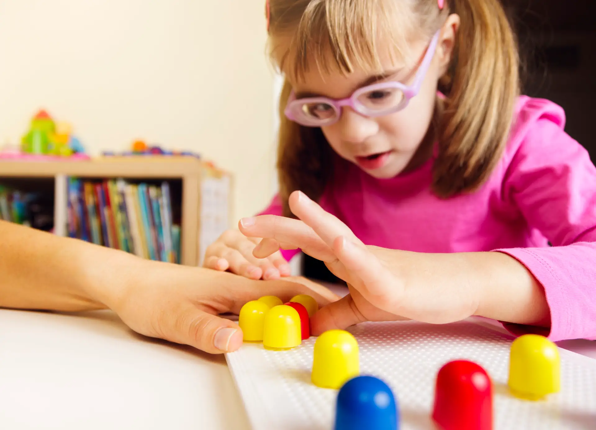 Young girl with low vision wearing a purple top and glasses, reaching for mosaic pegs on the board with an adult hand underneath her hand.