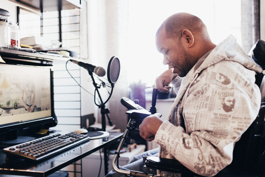 A man wearing a hoodie sitting in a power wheelchair faces a computer screen podcast equipment. He is looking at a joystick and smiling. His fingers are curled.