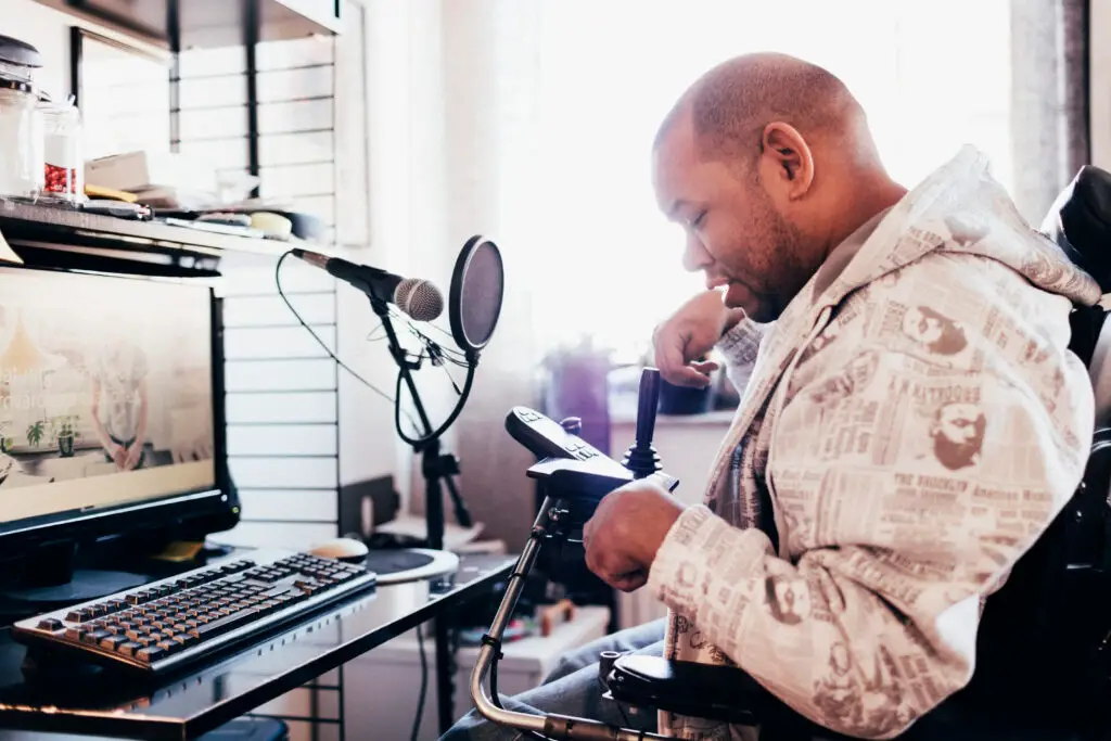 A man wearing a hoodie sitting in a power wheelchair faces a computer screen podcast equipment. He is looking at a joystick and smiling. His fingers are curled.