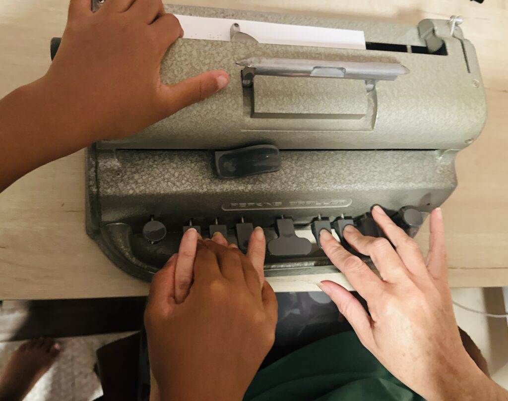 A trainer's hands type on a Perkins brailler. A young learner's right hand is placed over the instructors left hand and the left hand follows the braille on the paper.